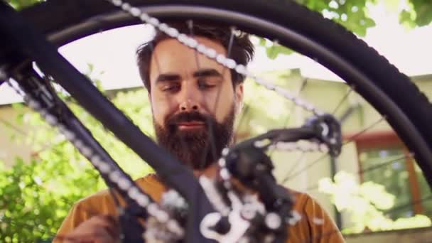 クローズアップショット 自転車の車輪が検査され 屋外の若い専用の慎重な男性によって修理されていることを示しています 庭の専門用具によって修理する損傷のための自転車タイヤを調べる健康な活動的な男 — ストック動画