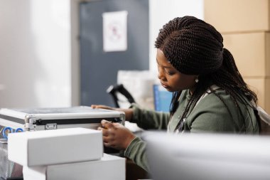 Metalik kutuya bakan depo yöneticisi, müşteriye göndermeden önce ürün kalitesini kontrol ediyor. Depo çalışanı tezgahta duruyor. Depoda paketleri hazırlıyor.