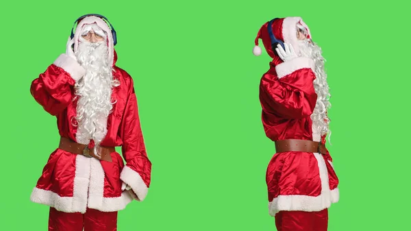 Modern man santa claus listen to music, dancing and having fun over greenscreen isolated backdrop. Young adult portraying saint nick in red costume enjoying songs on headset, celebration.