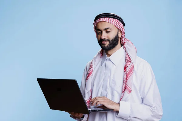 穆斯林男子身穿传统的正装 头戴头巾 手持笔记本电脑 在社交媒体上摇摆不定 阿拉伯人民穿着伊斯兰服装 使用手提电脑和查看网页 — 图库照片