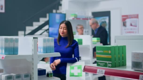 薬局の棚のなかで笑顔のアジアの顧客は 薬のパッケージを見て 問題を抱える病気のための適切な治療法を見つけようとしています ショッピングバッグを持ち 製品をブラウジングする楽しいコシェント — ストック動画