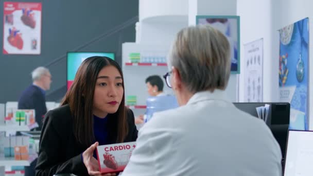 薬局のカウンターチェックアウト前のアジアの女性は 医療勧告のための資格のある薬剤師のキャッシャーを求めて 病気に対抗するために最高の医薬品を選択しようとしています — ストック動画