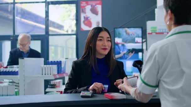 チェックアウトカウンターで麻薬店のアジア人女性は 医療品を購入するためにクレジットカードを使用しています 必要とされるウイルスの停止の丸薬を見つけた後薬局で無接触支払をする幸せな顧客 — ストック動画