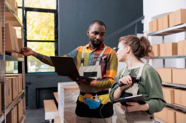Afro-Amerikalı yönetici ambarda ambalaj tarayıcısını kullanarak paketlerin barkodlarını tararken müşteri siparişiyle birlikte kutu gösteriyor. Ticaret envanterinde çalışan çeşitli çalışanlar