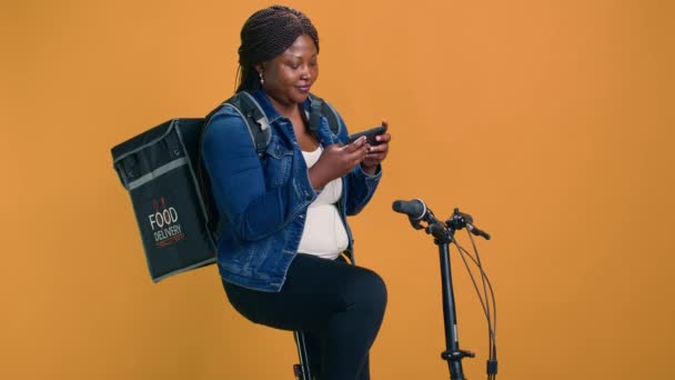 女信使休息时可以在自行车上使用智能手机放松自己 充满热情的非洲裔美国女人在等待网上信息传递的同时 还要检查手机 — 图库视频影像