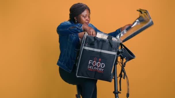 若いアフリカ系アメリカ人女性は 地元の顧客のための食品配達バッグからピザ箱を取り外しました オンデマンドで作業するクーリエは 効率的に輸送として自転車でファーストフードを提供します — ストック動画