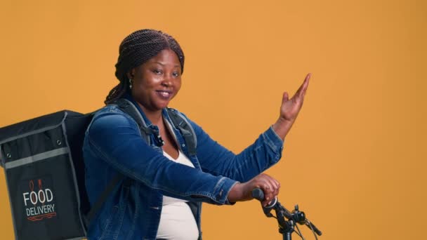 在黄色背景下 非洲裔美国妇女背着食物运送袋 自行车上的女骑手指向孤立的带有手部动作的彩色背景模板 — 图库视频影像