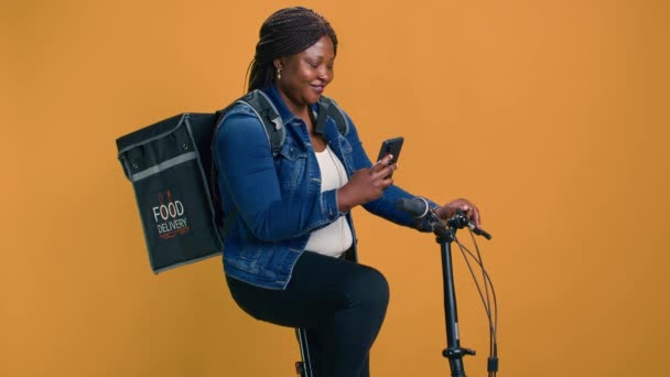 アフリカ系アメリカ人女性マルチタスクを自転車に乗せ 携帯電話で食べ物を届ける 若い黒人女性は 注文の効率的な輸送のためのモバイルデバイス上の配送アプリをチェック — ストック動画