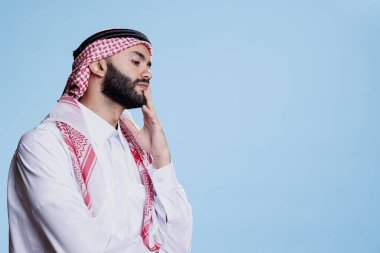 Geleneksel giysiler giyen düşünceli Müslüman adam stüdyoda düşünceli bir ifadeyle poz verirken çenesine dokunuyor. Kafası karışmış, kafası karışmış, çözümü düşünen Arap bir adam.