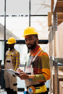 Afro-Amerikan lojistik danışmanı depo portresi envanteri çıkarıyor. Dağıtım yöneticisi ürün tedarik zinciri ve kamerayla ilgili panoya notlar alıyor