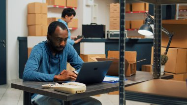 Male entrepreneur planning merchandise inventory and quality control, working on laptop to do retail logistics. Young man using stock in packages, shipping products and doing financial planning.