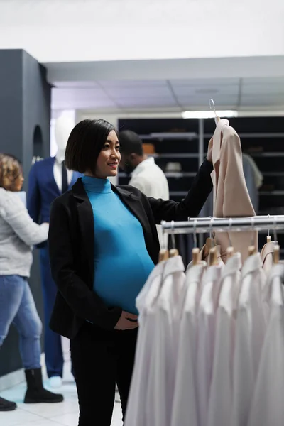 Smiling asian pregnant woman examining jacket on hanger while shopping for maternity clothes in mall. Expectant mother checking outfit on hanger while browsing rack with apparel