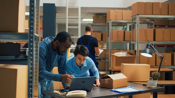 Offce employees doing teamwork to ship goods in cardboard boxes, checking quality of merchandise in storehouse space. Diverse owners using laptop to plan products shipment distribution.
