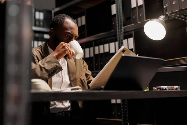 非洲裔美国警察在夜间喝咖啡和阅读Csi报告 侦探坐在办公室的办公桌前 拿着茶杯 在黑暗的办公室里研究犯罪案件档案 — 图库照片
