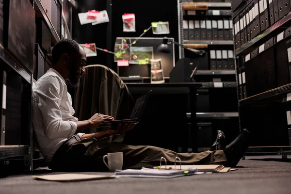Private investigation agency worker analyzing surveillance photos on laptop. African american detective sitting on floor and working late studying crime scene photographs on computer