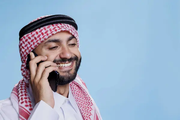 微笑的穆斯林男子穿着带着带绳索的头巾在手机上说话 穿着伊斯兰式头饰的快乐的阿拉伯人在智能手机上愉快地说话 — 图库照片