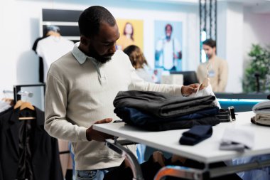 Afro-Amerikalı bir adam alışveriş merkezinde kıyafet alışverişi yapıyor. Mobil giyim mağazasının internet sitesine girmek için akıllı telefon kullanıyor. Uygulamada müşteri askıdan gömlek seçiyor ve mevcut stokları kaydırıyor