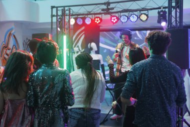 İnsanlar gece kulübünde dans edip eğlenirken, genç DJ sahne ışıkları altında sahne alıyor. Elektronik müzik konserinde kalabalık dans pistinde dans ediyor.