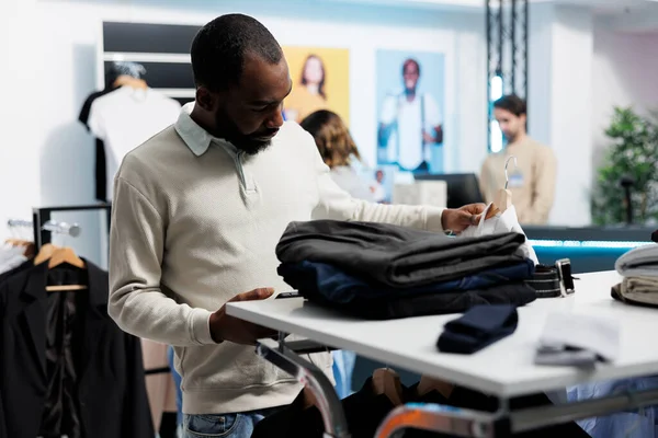 アフリカ系アメリカ人男性がモールでアパレルショッピングをしたり スマートフォンを使って服屋のモバイルサイトにアクセスしたりします ハンギングラックからシャツを選択し アプリで利用可能な在庫をスクロール — ストック写真