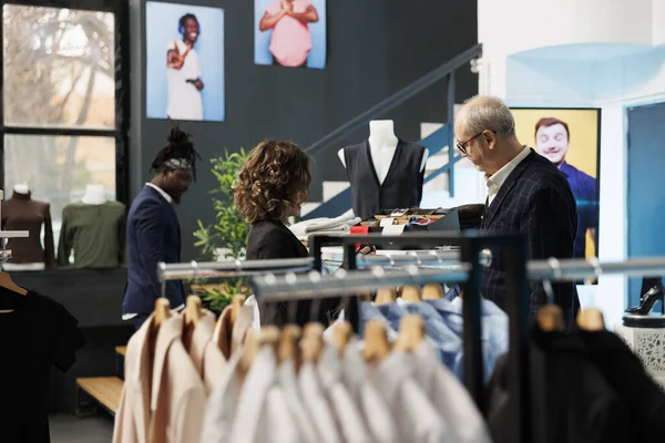 Senior man asking showroom worker for help with tie, customer shopping for formal wear in modern boutique. Elderly client buying fashionable clothes and stylish accessories. Fashion concept