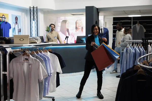 Clothing store cheerful smiling customer showcasing shopping bags with purchases and dancing. Happy excited asian woman carrying paper packages after buying apparel in boutique