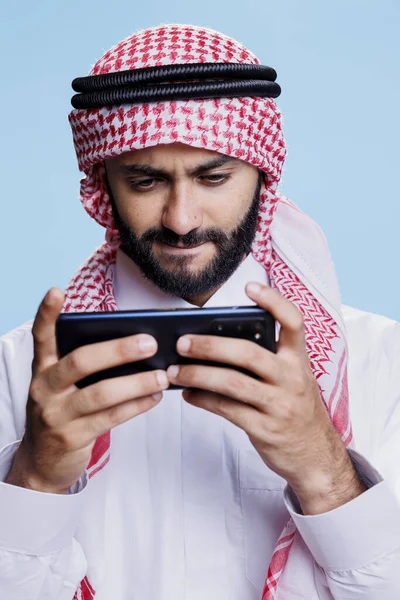 聚精会神的穆斯林男子头戴头巾 在智能手机上玩电子游戏 穿着传统服装 在手机上享受视频游戏应用的专注的Arab游戏玩家 — 图库照片