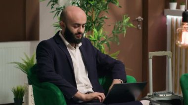 Orta Doğulu adam lobi alanında dinlenirken dizüstü bilgisayarda yazı yazıyor ve küresel şirket etkinliği için konuşma yazıyor. Profesyonel misafir iş gezisinde, otel resepsiyonunda..