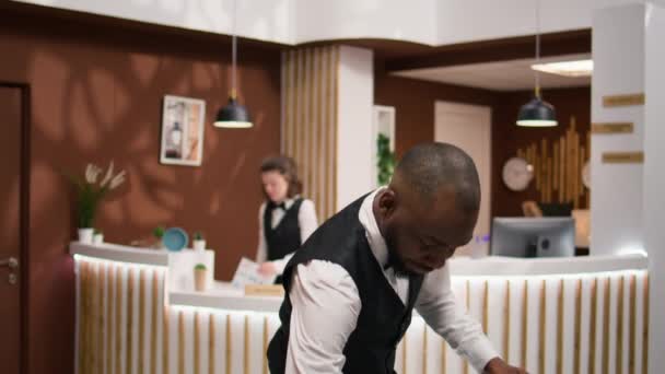 アフリカ系アメリカ人のベルボーイは ホテルのロビーで豪華なコンシェルジュサービスを提供する部屋の宿泊施設で重要なゲストを助けるためにスーツケースを取ります 従業員がビジネスマンになることと支援 — ストック動画