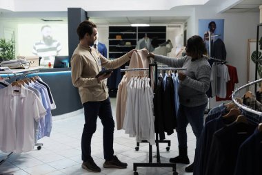 Moda butik asistanı rafları karıştırıyor ve müşteriye şık bir ceket sergiliyor. Elinde dijital tablet bulunan giyim mağazası çalışanı ve kıyafet seçmede danışman kadın alıcı.