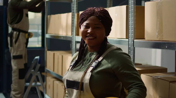 出荷する準備ができている段ボール箱の包装された商品で満たされた棚の前に立っている陽気なアフリカのアメリカの倉庫労働者の肖像画 専門の環境で働くこと幸せ — ストック写真