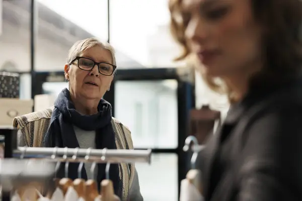 Selective focus of elderly customer asking employee for help with choosing shirt in clothing store. Senior woman shopping for casual wear, buying fashionable clothes in modern boutique