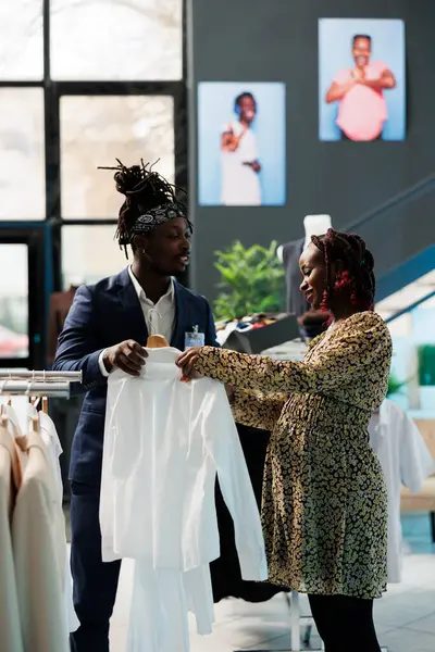 ショールームワーカーは 顧客が白いシャツを選択するのを支援し 衣料品店で服の生地を議論します アフリカ系アメリカ人妊婦がモダンなブティックでファッショナブルな母性商品を購入 — ストック写真
