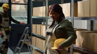 Karton kutularda paketlenmiş mallarla dolu rafların önünde duran neşeli Afro-Amerikan depo işçisinin portresi nakliyeye hazır, profesyonel çevrede çalışmaktan mutlu.