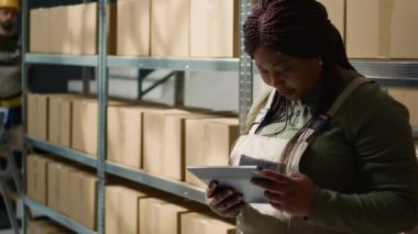 Neşeli Afro-Amerikan depo yöneticisinin portresi güncellenmiş ürün bilgilerini internete yüklüyor, perakende depolama tesislerinin arasında duruyor nakliyeye hazır paketleri tanımlıyor, kapatın.