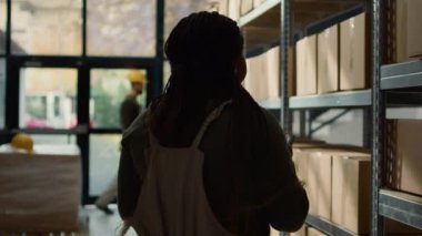 Envanter kontrol yöneticisinin görüntüsü güncellenmiş ürün bilgilerini çevrimiçi girerken, depolama sahalarının arasında durup malların etiketlerini incelerken stajyer de arka plandaki kutuları tarıyor.