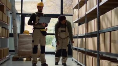 Çalışan ve Afrikalı Amerikalı iş arkadaşı depo koridorlarında yürüyor karton kutulardaki etiketleri kontrol ediyor, güncellenmiş ürünleri online alışveriş sitesine yüklüyor.