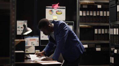 Odaklanmış Afro-Amerikan ajanı titizlikle kanıtları inceler, kayıtları araştırır ve raflardan ve laptoptan bilgi toplar. Siyahi polis ayakta duruyor ve ofis masasındaki dosyaları inceliyor..