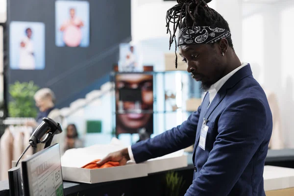 アフリカ系アメリカ人の従業員が現金登録に立って 顧客パッケージを準備しています ショールームワーカーは ショッピングセンターで働くカートンボックスにファッショナブルな服を置きます ファッションコンセプト — ストック写真