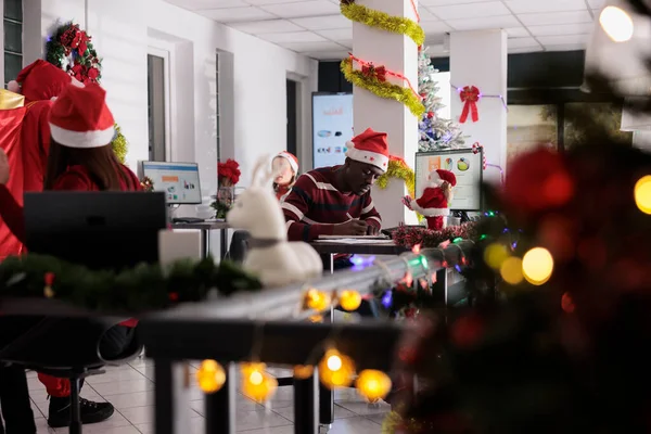 従業員は サンタクロースがお祝いの装飾オフィスでクリスマスプレゼントで彼を驚かすために来ることに気づいていません 冬の休暇中にオフィスに忍び込むXmasの衣装で偽装された労働者 — ストック写真