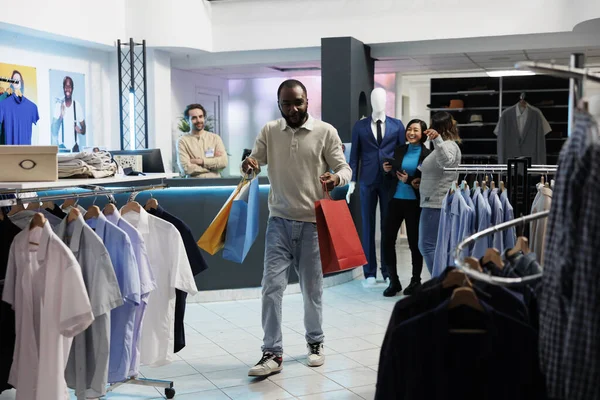 Happy shopaholic carrying paper bags filled with apparel in clothing store. Excited african american man holding packages while shopping and making purchase in mall boutique