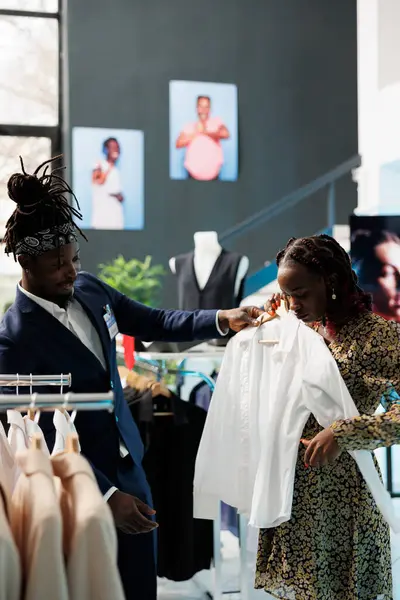 Boutique worker helping client with formal wear, looking at white shirt discussing material in clothing store. African american customer buying maternity clothes and trendy merchandise