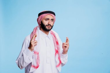 Kültürel Müslüman kıyafetleri giymiş korkmuş bir adam şans diliyor. Korkmuş, gözleri açık, stüdyoda mavi arka planda şans için batıl jestlerle poz veren bir Arap.
