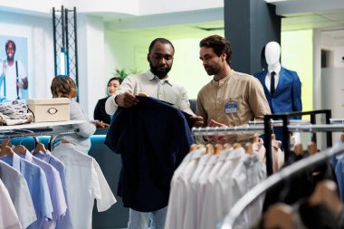 Afro-Amerikalı bir adam giyim mağazası çalışanlarına kıyafet gösteriyor ve moda tavsiyesi istiyor. Perakende pazarı butik asistanı ve müşteri şık gömleği birlikte seçiyor.