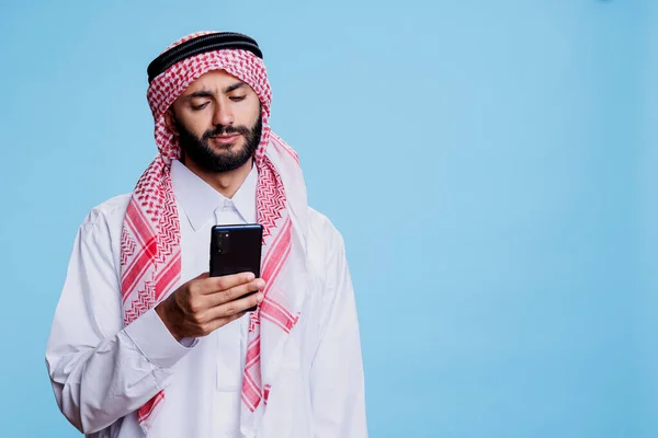 身穿传统服装的穆斯林男子手持智能手机 在工作室进行在线交流 阿拉伯人民穿着伊斯兰头巾和长袍 使用手机社交媒体 — 图库照片