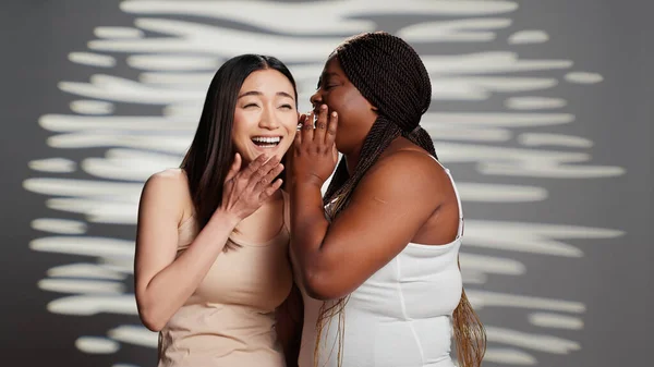 African american woman whispering secret to asian girl on camera, acting sensual and glamorous in studio. Interracial skincare models promoting body positivity and acceptance, having fun.