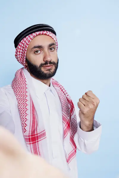 ギュラートのヘッドドレスを身に着けている真剣なイスラム教徒の男性は ひび割れた拳を見せて スマートフォンのフロントカメラを見ています 携帯電話の貧困を抱えながら積極的なジェスチャーを作るアラブ — ストック写真