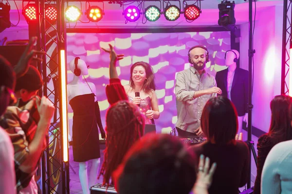 Man and woman musicians playing and singing on stage at electronic music festival in nightclub. Singer and dj techno band performing while people dancing on dancefloor in club