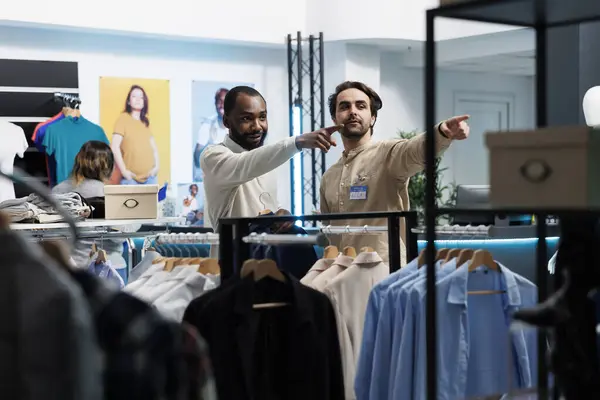 アフリカ系アメリカ人男性がラックを指差し 衣料品店のアシスタントにアパレルサイズのオプションについて尋ねました ショッピングセンターのブティック顧客がコンサルタントの支援を受ける — ストック写真