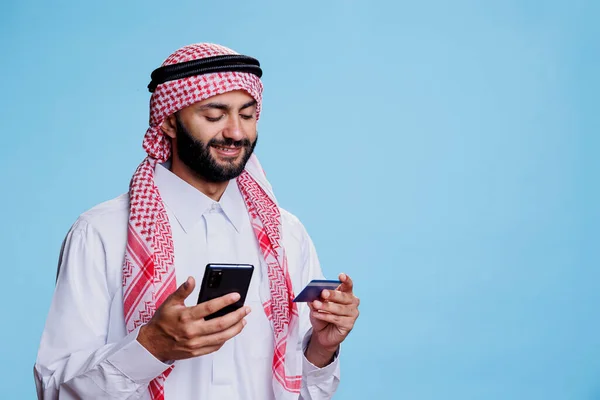 用借记卡在网上商店用智能手机购物时微笑的穆斯林男子 在手机网站上购买伊斯兰头巾和头巾的阿拉伯人 — 图库照片