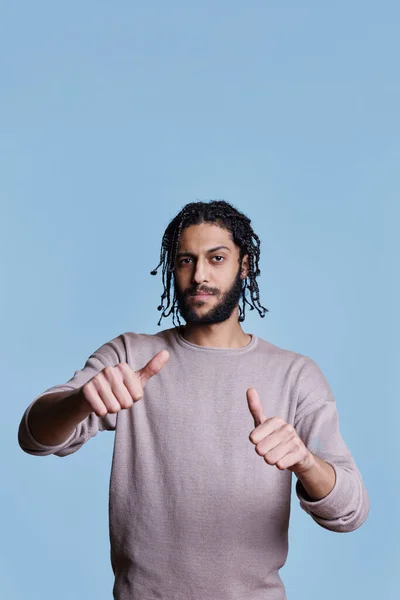 クールなアラブの男はジェスチャーの上に2つの親指を示し 眉を上げたカメラを見ています 自信を持って本格的な大人の人が指の肖像画でシンボルのように作る — ストック写真